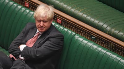 Boris Johnson sitter ensam med korslagda armar på en av de gröna bänkarna i parlamentet. 