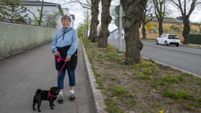 En kvinna i glasögon står med sin hund på en trottoar och tittar in i kameran.