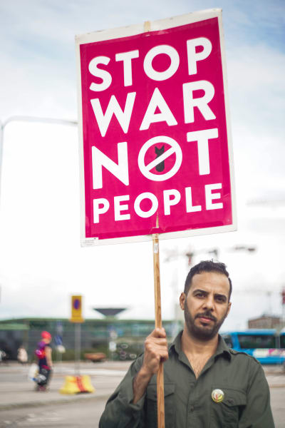 Saad Ali står på ett torg vid Hertonäs metrostation. Han har en skylt i handen, som det står "Stop war not people" på.