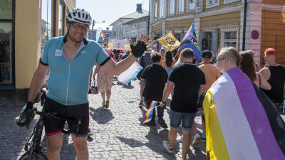 Mikael Kokkola står med sin cykel och vinkar, bredvid honom går ett pridetåg med mängder av människor.
