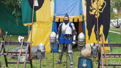 Haarniskaan pukeutunut mies seisoo keltasinisen teltan edustalla, edessään erilaisia keskiaikaisia aseita ja kypäriä.
