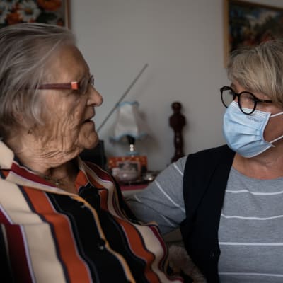 Närvårdaren Manuela Nordström, klädd i grått med svart väst och munskydd, talar med Gretel Gestrin, i randig blus