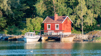 en röd stuga vid vattnet med en båt förtöjd vid en liten brygga. Mycket skog bakom.