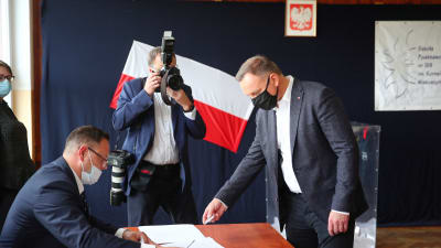 President Andrzej Duda röstar i presidentvalet i Polen den 12 juli 2020. En man ger en valsedel till en annan man, båda bär munskydd. En tredje man fotograferar.