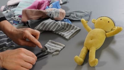 Hand som klipper till ett par docktrosor av en strumpa. En docka ligger också på bordet samt en hög med gamla strumpor.