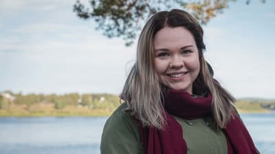 Nainen viininpunainen huivi kaulassa ja vihreä hupputakki päällä hymyilee ja katsoo kameraan, taustalla jokimaisema