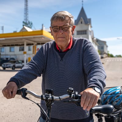 Äldre man leder sin elcykel och ser mot kameran, i bakgrunden en bensinmack i centrum av Hangö.
