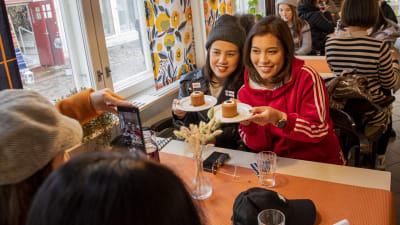 Två thailändska kvinnor sitter i ett café. De poserar med två Runebergstårtor på ett fat och en kvinna som sitter mitt emot tar bilden med sin telefon.
