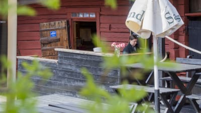 En kvinna jobbar vid en bardisk vid en uteservering. I förgrunden syns ett parasoll och några gröna blad.