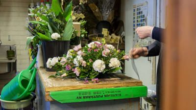 En blomsteruppsättning för en begravning ligger på ett bord i en blomsterhandel. Till höger syns en i personalen som håller på och gör en rosett.