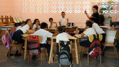 Pieni koululaisryhmä kuuntelee opettajaa luokkahuoneessa, heidän pulpettinsa on aseteltu ympyrään.