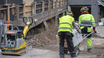 Två byggarbetare transporterar betongelement med en kärra på ett bygge