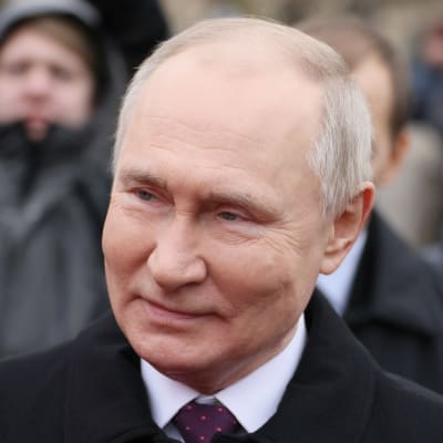 Närbild av Vladimir Putin som småler och tittar åt sidan. I bakgrunden syns människor utomhus.