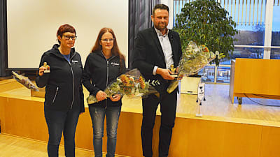 två kvinnor och en man med blommor i handen på prisutdelning.