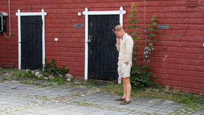 En man i ljusa kläder står utomhus framör en röd trävägg. Han talar i telefon och tittar ner i marken.