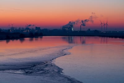 Anläggningar för fossilt bränsle och vindkraftverk vid vatten i solnedgång.