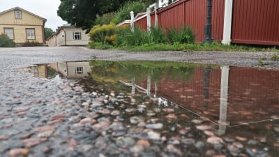 Bild av en vattenpöl på asfalterad yta. I vattenpölen speglas ett plank och några hus som ligger i bakgrunden. Vattenpölen i fokus. 