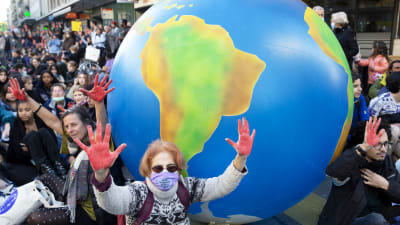 Personer i klimatdemonstration. En äldre kvinna med solglasögon och munskydd står framför en uppblåst jordglob och med uppsträckta händer.
