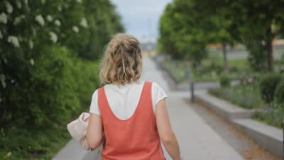 Vit kvinna med ljust hår går med ryggen mot kameran på en gångväg med gröna träd runt.