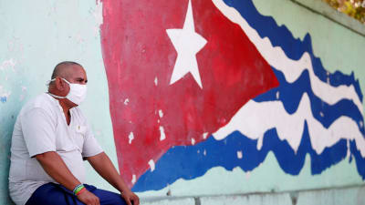 Man med munskydd invid den kubanska flaggan. Havanna, Kuba 21.4.2020