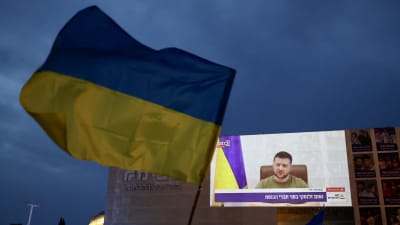 I förgrunden en ukrainsk flagga som vajar i luften. I bakgrunden Ukrainas president Volodymyr Zelenskyj som syns på en tv-skärm på en byggnads vägg.