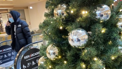 Bilden är tagen i ett köpcenter. Närbild på en julgran med lampor och silvriga stora julgransbollar. I bakgrunden ser man en rulltrappa som andänds av en person med uppdragen huva och munskydd.