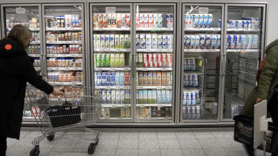 Kunder vid mjölkhyllan i en affär. Bakom glasdörrar finns hyllvis med olika mjölkprodukter. Till höger i kallskåpet syns metallkärror med mjölkburkar på.