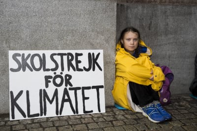 Greta Thunberg i en gul regnrock sitter på marken bredvid en skylt där det står "skolstrejk för klimatet".