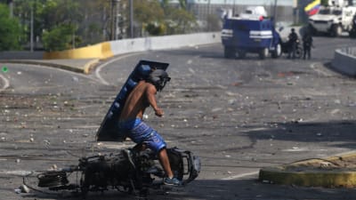 En sköldförsedd demonstrant springer förbi en utbränd motorcykel under kravellerna i Caracas 