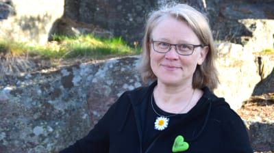Blond kvinna stå vid ett berg. Hon är klädd i svart och har ett smycke som föreställer en prästkrage.