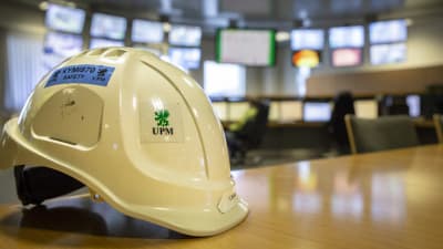 En skyddshjälm på ett bord i ett industriellt kontrollrum med många skärmar i bakgrunden.  