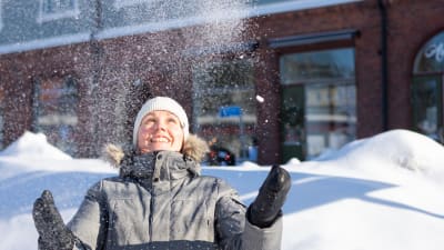 En vinterklädd kvinna ser glad ut och kastar upp snö i luften, snöhögar, trafikmärken och hus i bakgrunden.