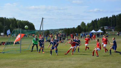 Två pojklag, i röda och i blå skjortor, försöker nå ett inlägg i straffområdet under en fotbollsmatch.