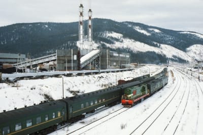 Tåg på en station bredvid en industrianläggning i bakgrunden syns höga berg