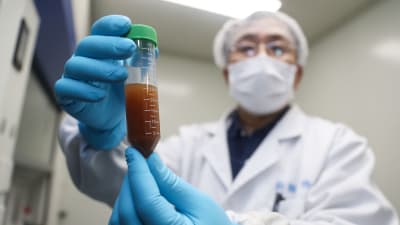 En person i skyddmask och vit läkarrock visar upp en liten medicinflaska. 