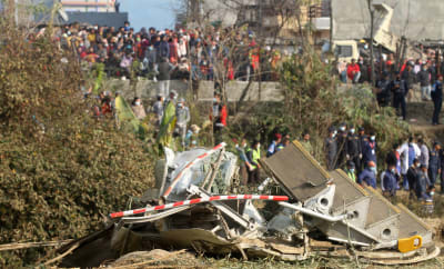 Bråte från ett flygplan ligger på marken, i bakgrunden syns grupper med människor.