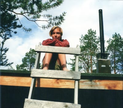 Riikka Suominen istuu teininä mökin katolla. Etualalla tikkaat, Riikka nojaa tikkaisiin ja katsoo kameraan.
