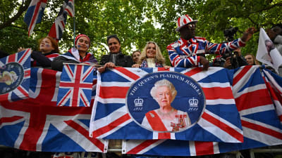 En rad människor med brittiska flaggor och ett porträtt av drottning Elizabeth framför sig. I bakgrunden lövträd.