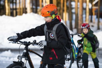 Två barn i vinterkläder och cykelhjälm leder sina cyklar över en snöig skolgård.