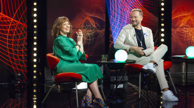 Två personer som sitter i en studio, en klädd i grönt och en i en ljus kostym. De ser på varandra och ser glada ut.