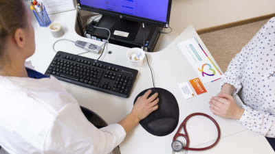 En läkare sitter vid datorn och skriver och mittemot sitter en patient.