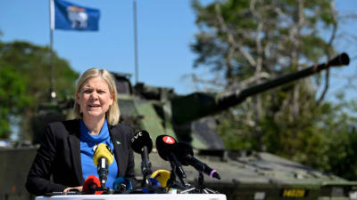 Magdalena Andersson med mikrofoner framför sig. I bakgrunden en pansarvagn och en blå flagga.