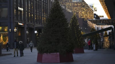 Två granar med julljus står i förgrunden, på gatan går vinterklädda människor. På byggnaderna hänger julljus.