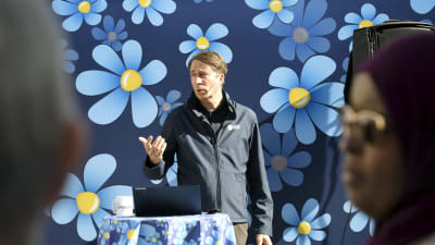 Richard Jomshof talar med en sverigedemokratiskt blommig bakgrund bakom sig.