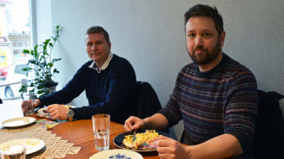 Två män sitter vid ett bord med mat framför sig.