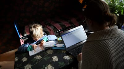 En pojke sitter och gör skolarbete vid ett bord. På andra sidan bordet sitter en kvinna och jobbar på en bärbar dator.