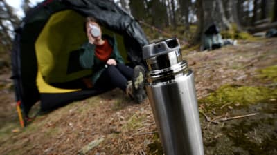 En kvinna sitter halvt inne i ett tält i skogen och för en mugg till munnen. I förgrunden en öppnad termos.