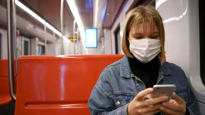 En ung kvinna sitter i metron i Helsingfors. Hon bär munskydd och tittar ner på sin mobil.