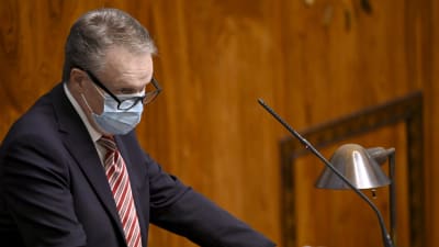 Ilkka Kanerva står i en talarstol i riksdagen. Han har munskydd på sig.
