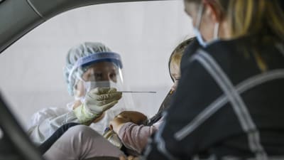 En sjukskötare i skyddsutrustning testar ett barn för coronaviruset. Barnet sitter i en bil i en bilstol, bredvid henne sitter en vuxen kvinna i munskydd.
